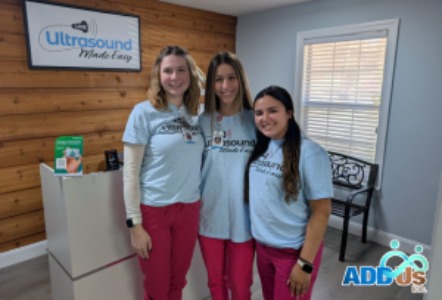 Three women wearing matching “Ultrasound Made Easy” shirts stand together smiling inside a clinic space, with a sign for the organization visible on the wall behind them.