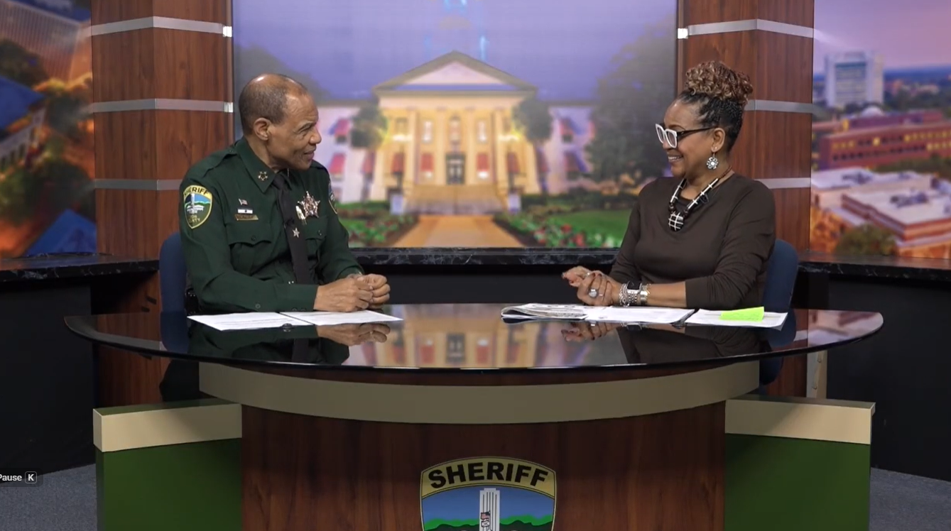 Two people seated at a news-style desk in a studio, including a uniformed sheriff and a host, engaged in conversation with the Florida State Capitol building displayed on the backdrop.